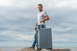 © yaroslav1986 - Young man in jeans and a white shirt stands with a gray suitcase on the seashore on huge rocks against a sky with clouds and looks into the camera
