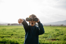 Surprised Boy Pointing While Looking Through Binoculars In Field During Sunny Day