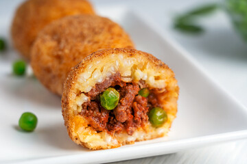 Wall Mural - Close up view of homemade Arancini italian rice balls, coated with bread crumbs, deep fried and stuffed with minced meat and green peas served on rectangular plate on white wooden background