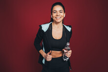 Smiling Sportswoman In Sports Clothing Holding Water Bottle Against Maroon Background