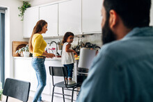 Father Looking At Daughter Helping Mother In Kitchen