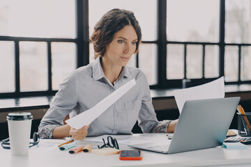 Focused hispanic woman worker analyzing documents and working on laptop computer in office