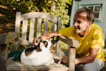 Smiling Man Crouching While Stroking Fluffy Cat Sitting On Armchair In Back Yard