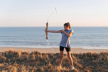 Archeress Aiming With Bow And Arrow While Standing On Grass At Beach
