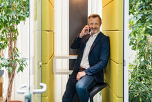 Smiling Businessman Talking On Smart Phone While Sitting In Soundproof Booth
