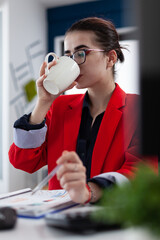 Poster - Manager in red jacket at startup desk drinking from white cup. Employee with glasses having coffe or tea break at the office. Businesswoman taking a pause from reading chart on clipboard.