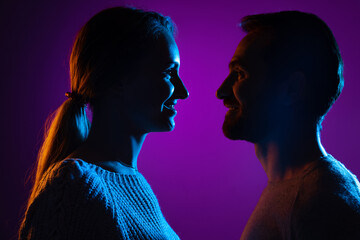 Cropped portrait of young couple standing face to face to each other isolated over purple background in neon lights