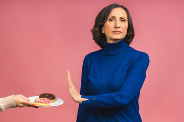 Stop sign. Charming happy aged mature senior woman refused to eat glazed donut dressed in casual isolated over pink background. Old age and diet concept.