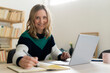 © Westend61 - Blond woman smiling while sitting by laptop on table at home