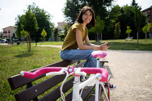 Smiling Woman Sitting On Bench Near Bicycle On Footpath