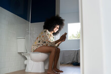 Smiling Woman Using Smart Phone While Sitting On Toilet Seat At Home