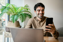 Smiling Male Freelancer Using Mobile Phone While Sitting With Laptop At Cafe