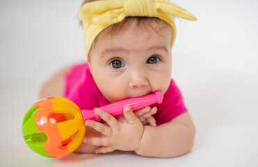 Wall Mural - cute, beautiful little girl in a pink bodysuit
 and a bandage lies on a white background, smiles and gnaws toys. beautiful baby. teeth are erupting. portrait of a baby. portrait of a beautiful girl