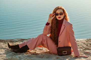 Fashionable woman wearing classic pink suit blazer, trousers, suede boots, trendy sunglasses, with small leather bag, posing on sandy coast. Copy, empty space for text