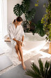 Smiling young woman in bathrobe dry brushing her legs with a natural wooden brush while sitting on the side of a bathtub in the bathroom decorated with green plants. Skin and body care, spa concept