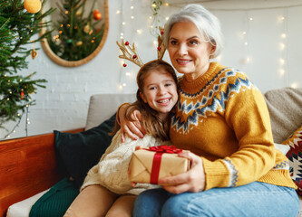 Sticker - Little girl granddaughter giving Christmas gift box to smiling grandmother during winter holidays