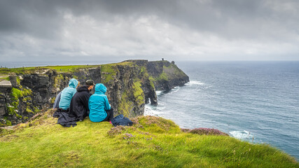 Group of young people sitting on the edge of cliff on iconic Cliffs of Moher, popular tourist attraction, Wild Atlantic Way, County Clare, Ireland