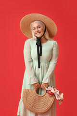 Poster - Mature woman in straw hat holding bag with bouquet of beautiful flowers on red background