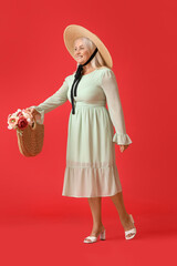 Poster - Beautiful mature woman in straw hat holding bag with bouquet of flowers on red background