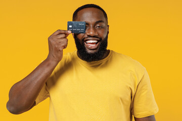 Wall Mural - Young smiling satisfied fun rich cool happy black man 20s wear bright casual t-shirt cover eye with credit bank card isolated on plain yellow color background studio portrait People lifestyle concept.