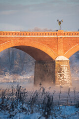 Wall Mural - Long red brick bridge over Venta river in misty, sunny, snowy winter morning with frosted trees, Kuldiga, Latvia