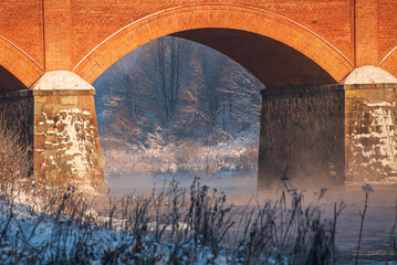 Wall Mural - Long red brick bridge over Venta river in misty, sunny, snowy winter morning with frosted trees, Kuldiga, Latvia