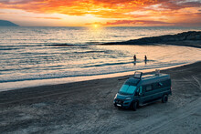 Aerial Photo Of Campervan On Abandoned Beach Against Beutiful Sunset. People Bathing In The Sparkling Sea. Outdoor Nomad Lifestyle, Van Life Holiday. Independent Road Trip Concept.