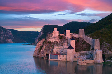 Wall Mural - Aerial, across lake view of the medieval fortress Golubac over  Danube river. Fortress towers illuminated by pink light. Sunset, pink and red clouds sky. Outdoor and traveling castle theme. Serbia.