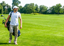 Young Male Golfer Carries A Bag Of Golf Clubs. Slim Man In White T-shirt , Golf Shoes, White Cap And Beige Pants Is Going In The Green Turf Grass. Copy Space. Full-length