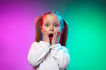 Studio shot of shocked, scared little girl, kid standing with open mouth isolated on gradient purple blue color background in neon light