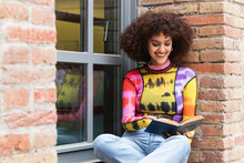 Smiling Curly Hair Woman Reading Book While Sitting On Window Sill