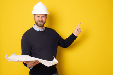 Young architect man wearing contractor helmet holding project paper plan pointing finger up over isolated yellow background