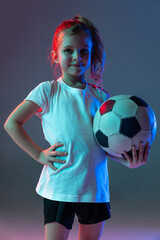 Wall Mural - Cropped portrait of little girl, posing with football ball isolated over gradient blue background in neon lights
