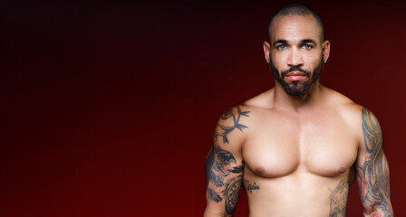 Wall Mural - Man of 30-40 years old with beautiful muscular body with tattoos stands on red dark background and looks directly into camera. Advertising banner with space for copying. Studio portrait.