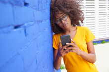 Afro Woman Using Mobile Phone While Leaning On Blue Wall