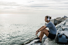 Sportsman With Headphones Looking At View While Sitting On Rock By Sea