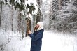 © Natalya - Young attractive woman with long hair, blue eyes, dressed in blue coat, white hat decorates Christmas tree in beautiful winter snow forest