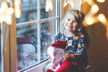 Little preschool girl holding cup Santa Claus boot with gift called Nikolausstiefel in German. Happy child wait on holiday by window with Christmas lights in winter. Cozy family celebration of xmas.