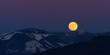 A winter wolf moon touches a mountain ridge with patches of snow where trees have been clearcut.  The moon is pink from the recent sunset and the sky is purple