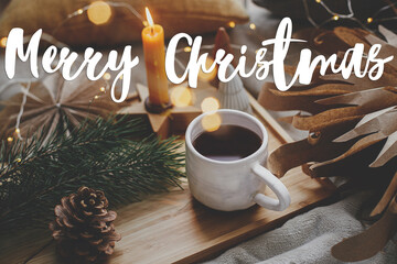 Wall Mural - Merry Christmas text on warm cup of tea, christmas lights, stars, pine trees, candle. Winter hygge. Season's greetings card. Handwritten sign. Happy Holidays!