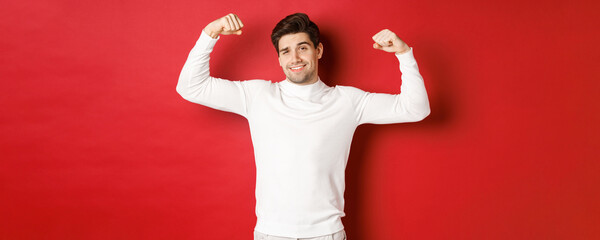 Portrait of smiling handsome man in white sweater, flexing biceps and bragging with strength, show-off strong muscles after workout, standing over red background