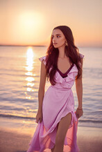 Portrait Of A Beautiful Young Caucasian Girl Walking Along The Beach With A Nice Pink Sunset And Purple Water In The Background.