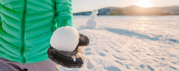 Winter snowball fight concept outdoors. Girl holding snowball on snowy day with snowman in background on frozen lake at beautiful winter sunset living active lifestyle. Panoramic banner