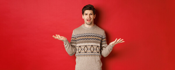 Portrait of confused and disappointed handsome guy, complaining about christmas, spread hands sideways and frowning displeased, standing in xmas sweater over red background