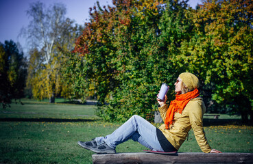 Wall Mural - Happy woman enjoying coffee on park bench on green foliage background. Autumn concept. Leisure time in sunny fall day. Copy space.