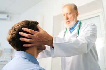 Close-up low-angle view of mature adult male doctor massaging temples to sick patient during consultation at hospital. Young man with headache having help during checkup visit in clinic office.