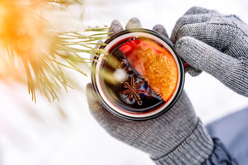 Mulled wine in the hands of a girl during a snowfall in the forest. Winter hot drinks with aromatic spices of cinnamon, cardamom and orange. Warmth, comfort and atmosphere of December nature and