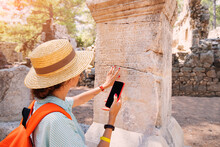 Woman Philologist And Tourist With Smartphone Reads And Translates Ancient Greek From Columns In The Ruins Of An Antique City. Linguistics And Archaeology