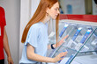 © alfa27 - Side View On Female Interested In Smartphone In Store, Make Purchase, Redhead Lady With Long Hair In Casual Wear Looking At Mobile Phone Through Glass. Discounts In Tech Shop.