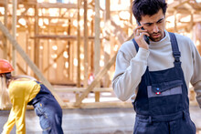 Upset Builder Architect Man Talking On Phone Against New Residential Construction Home Framing, At Sunny Day Outdoors, Wearing Working Uniform, Female Construction Companion In The Background.